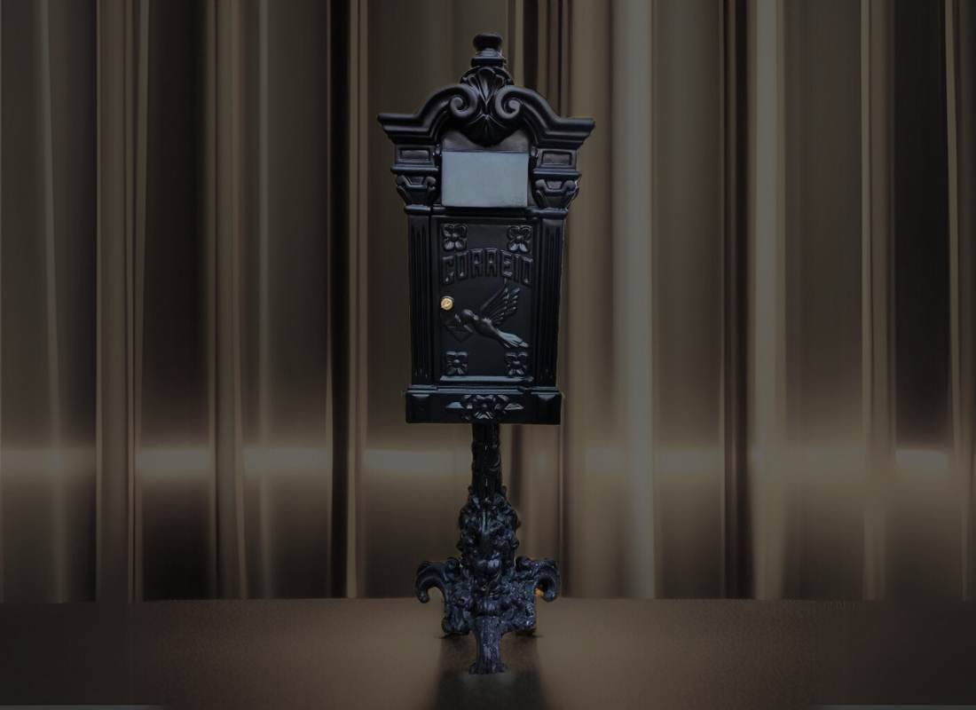 Caixa de correio preta em estilo clássico, com ornamentos, a palavra Correio e um pássaro em relevo, sobre pedestal decorado