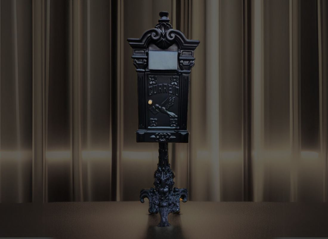 Caixa de correio preta em estilo clássico, com ornamentos, a palavra Correio e um pássaro em relevo, sobre pedestal decorado